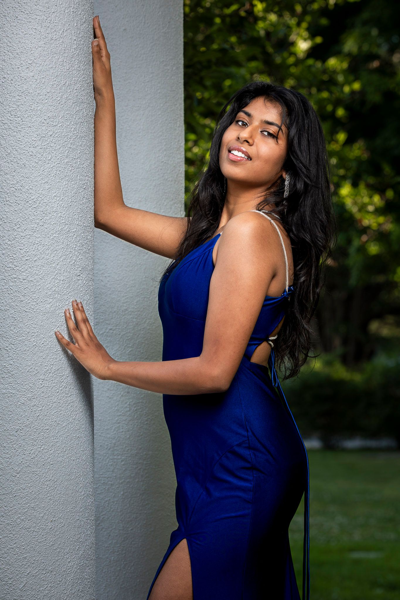 Young woman in royal blue satin gown with slit poses on outdoor deck, smiling elegantly.