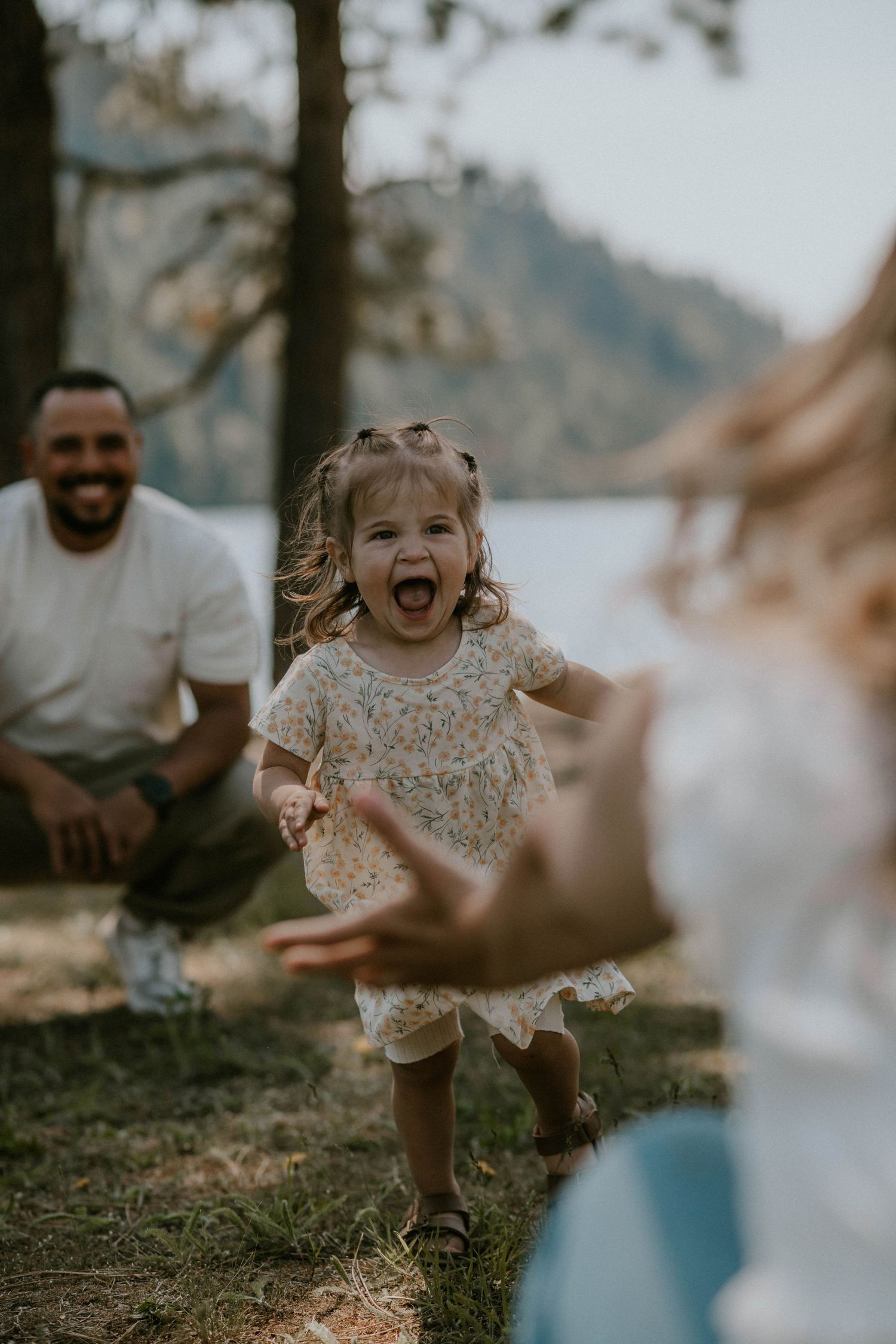 A couple smiles warmly at their toddler daughter playing outdoors near a lake surrounded by trees.