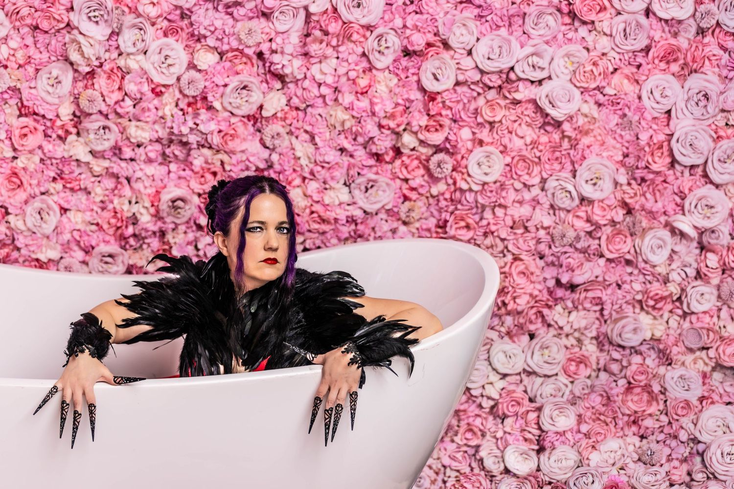A goth woman in black feathers and long black fingernails sits inside a white bathtub in front of a wall of pink flowers.