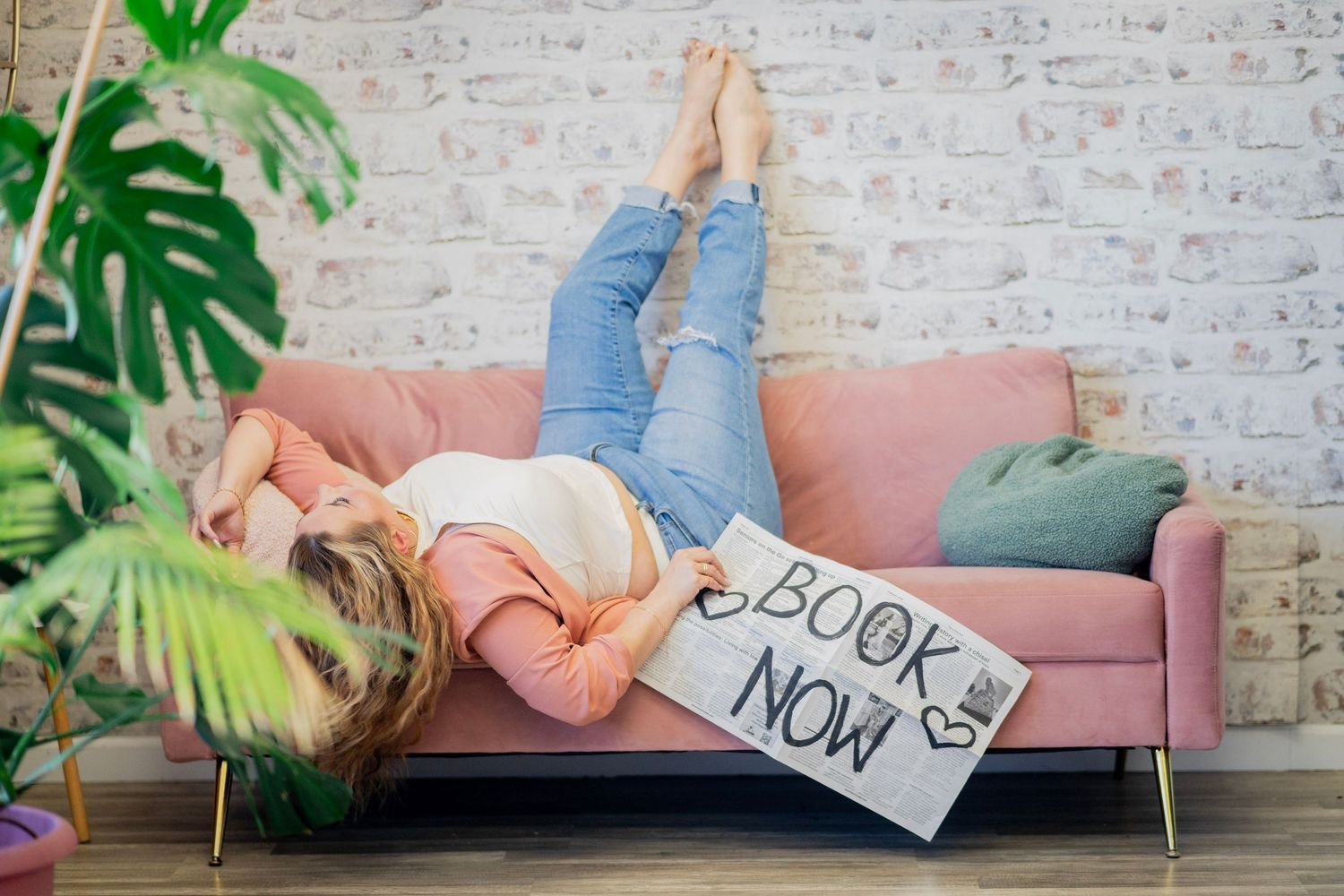 Woman lying on a pink sofa holding a Book Now sign with legs up against a white brick wall.