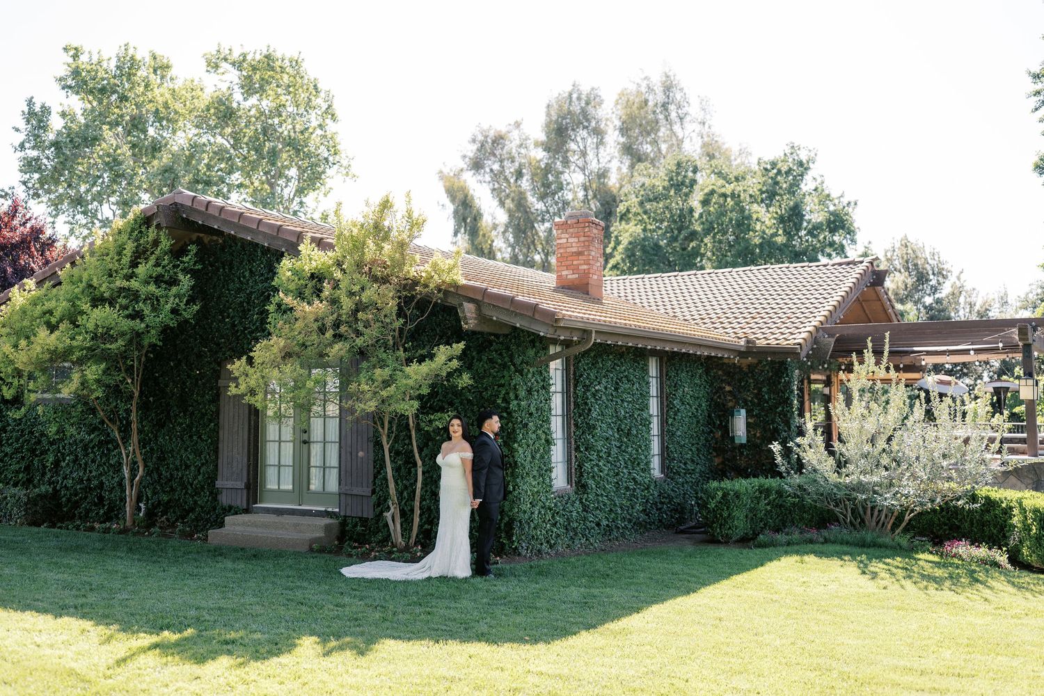 Bride and groom pose in front of an ivy-covered brick wedding venue surrounded by lush green lawns and trees.