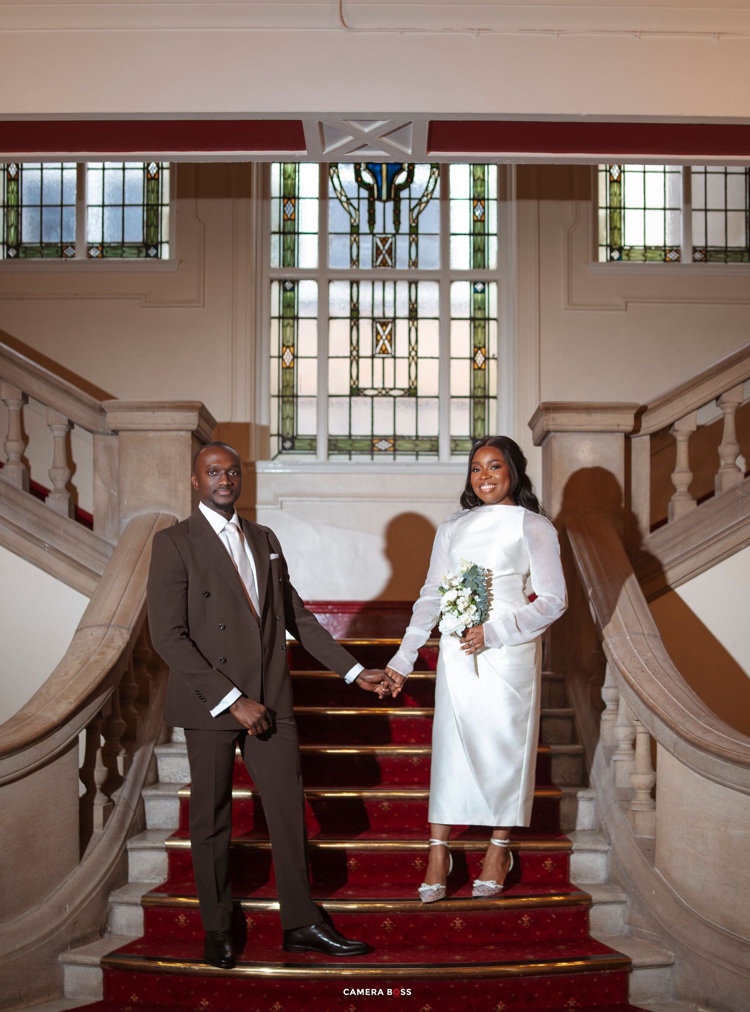 A couple in wedding attire holds hands on a grand staircase with red carpet and stained glass window backdrop.