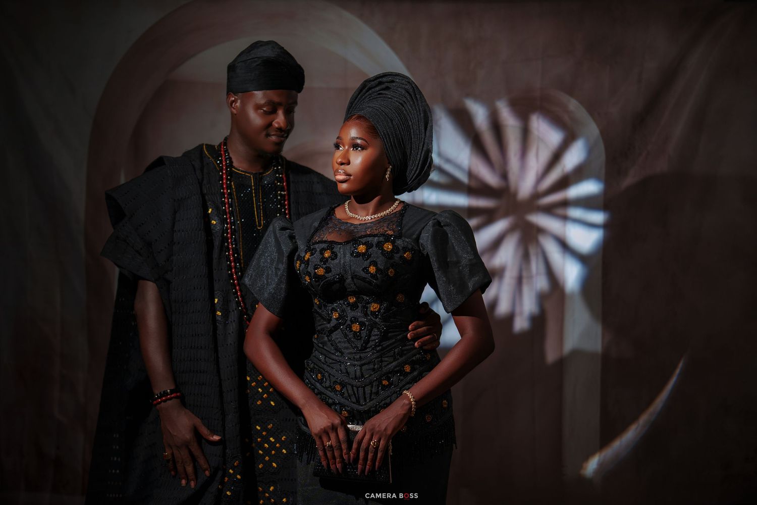 A couple in elegant black traditional African attire poses for a pre-wedding portrait against a dramatic shadowed backdrop.