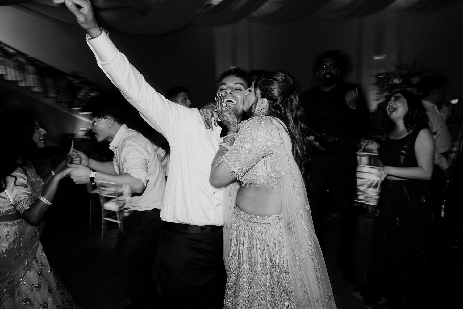Black and white photo of a couple dancing joyfully at an Indian wedding reception surrounded by guests.