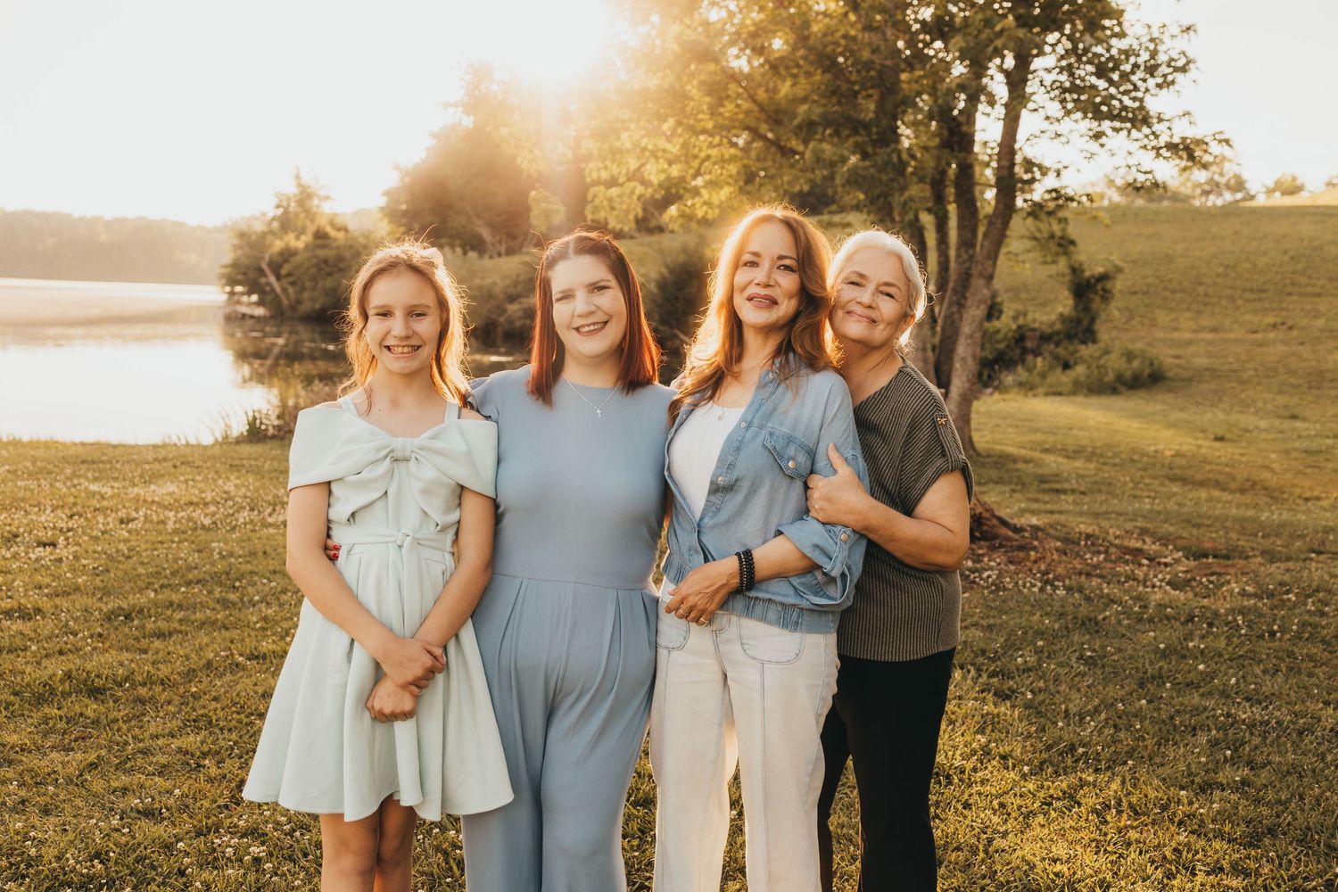 Four women smiling together outdoors at golden hour near a lake, wearing casual outfits in soft blue and neutral tones.