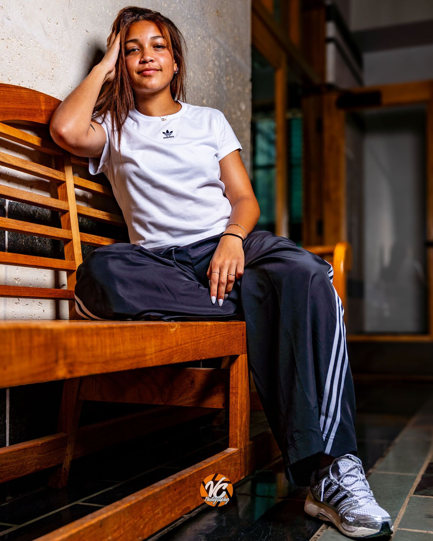 Young woman in white Adidas t-shirt and navy track pants sitting relaxed on a wooden bench.
