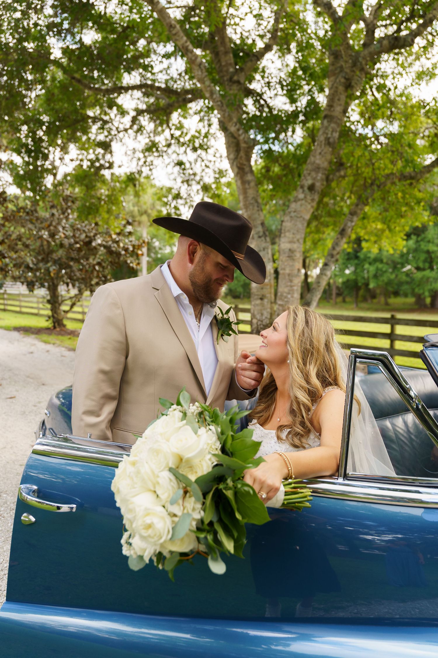 Groom in cowboy hat kisses bride's hand beside a classic blue convertible, holding white bouquet outdoors.