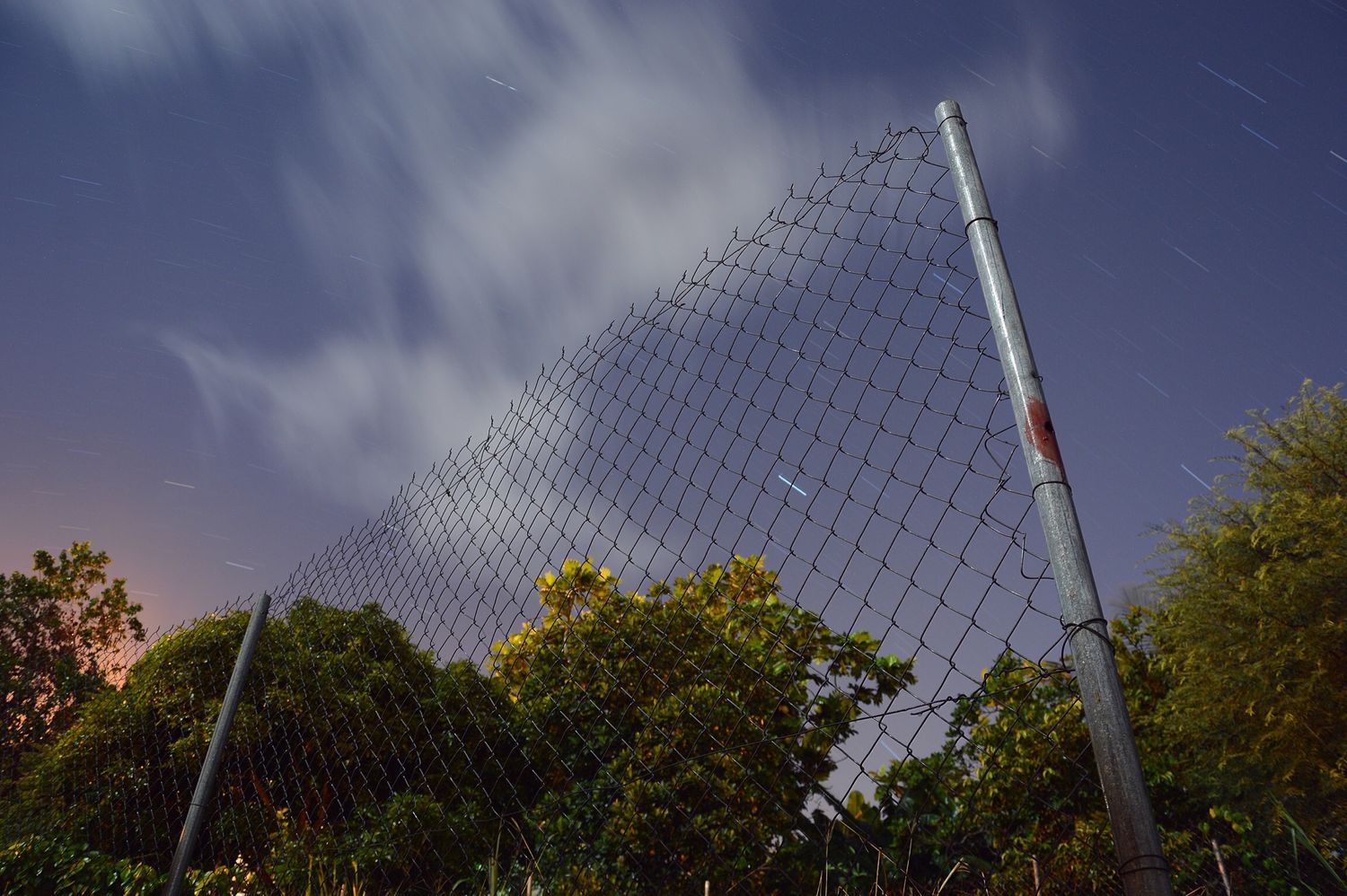 Chain-link fence stretching upward against a dramatic night sky with stars and clouds, surrounded by lush green trees.