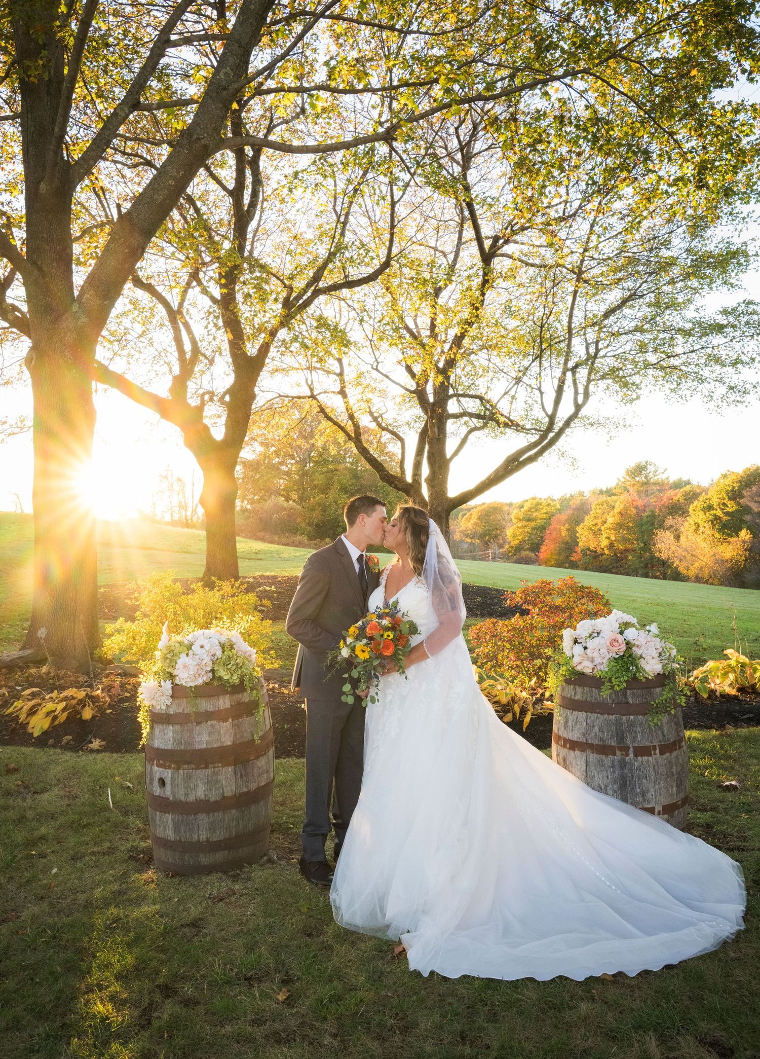 Bride and groom share a kiss at golden hour on a sunlit lawn, framed by autumn trees, floral-topped barrels, and a flowing wedding gown.