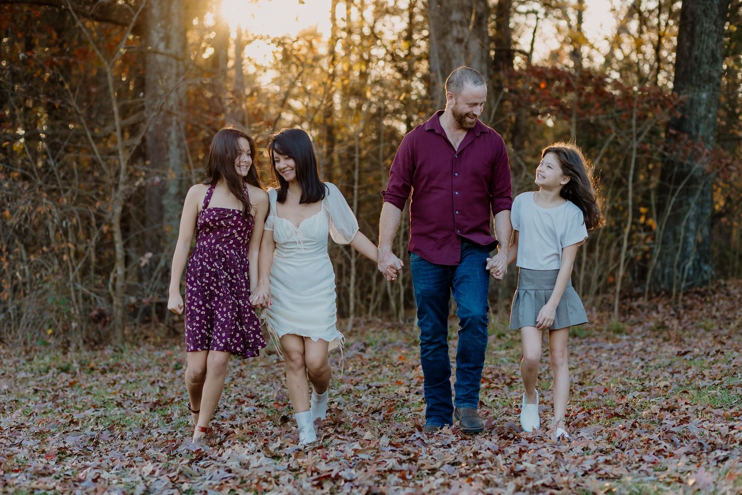 Family of four walking hand-in-hand through autumn woods at golden hour, smiling and enjoying a peaceful outdoor stroll.