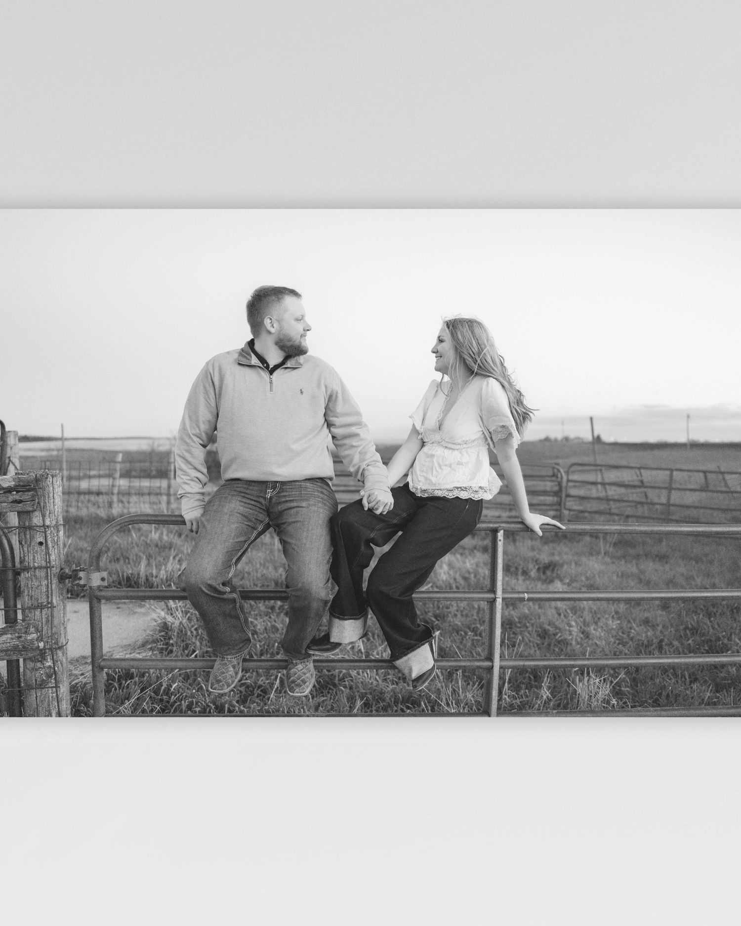 Black and white photo of a couple sitting on a farm gate holding hands and smiling at each other in a rural field.