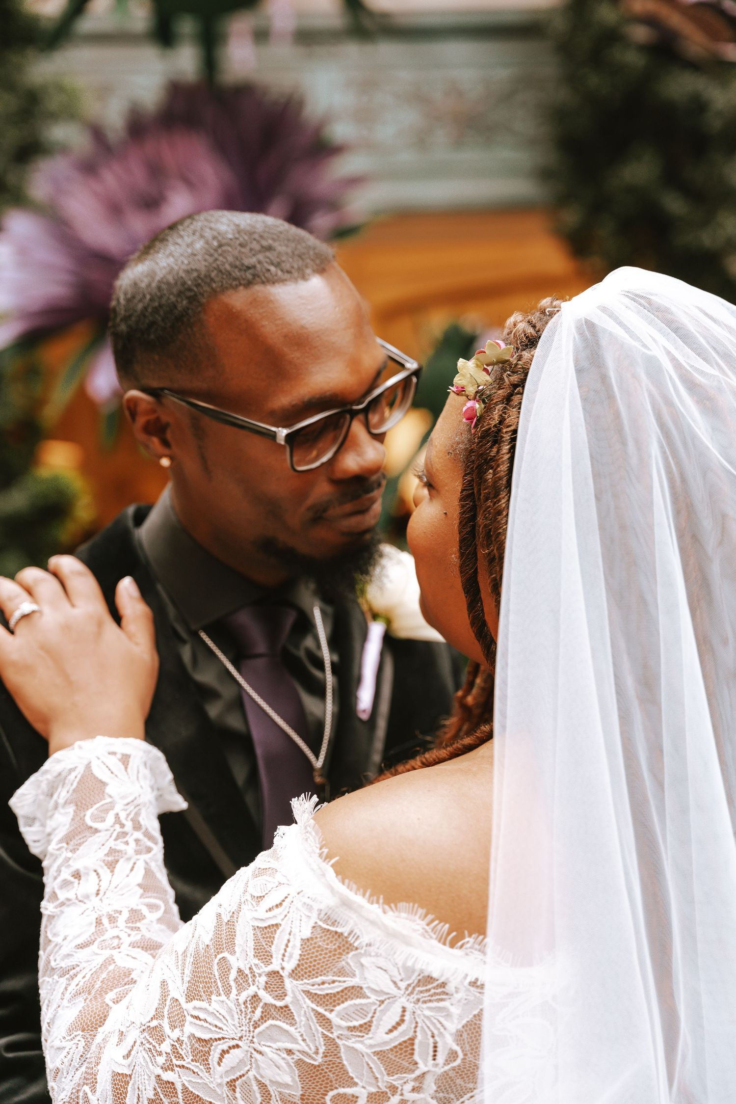 A smiling bride and groom pose together in a lush garden venue, bride wearing white gown and veil, groom in black suit.