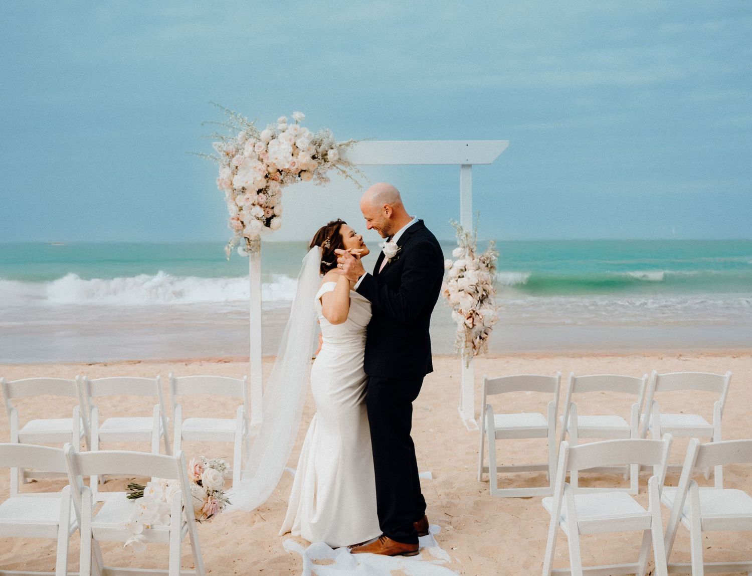 Couple sharing a romantic moment at a beach wedding ceremony with floral arch and ocean backdrop.