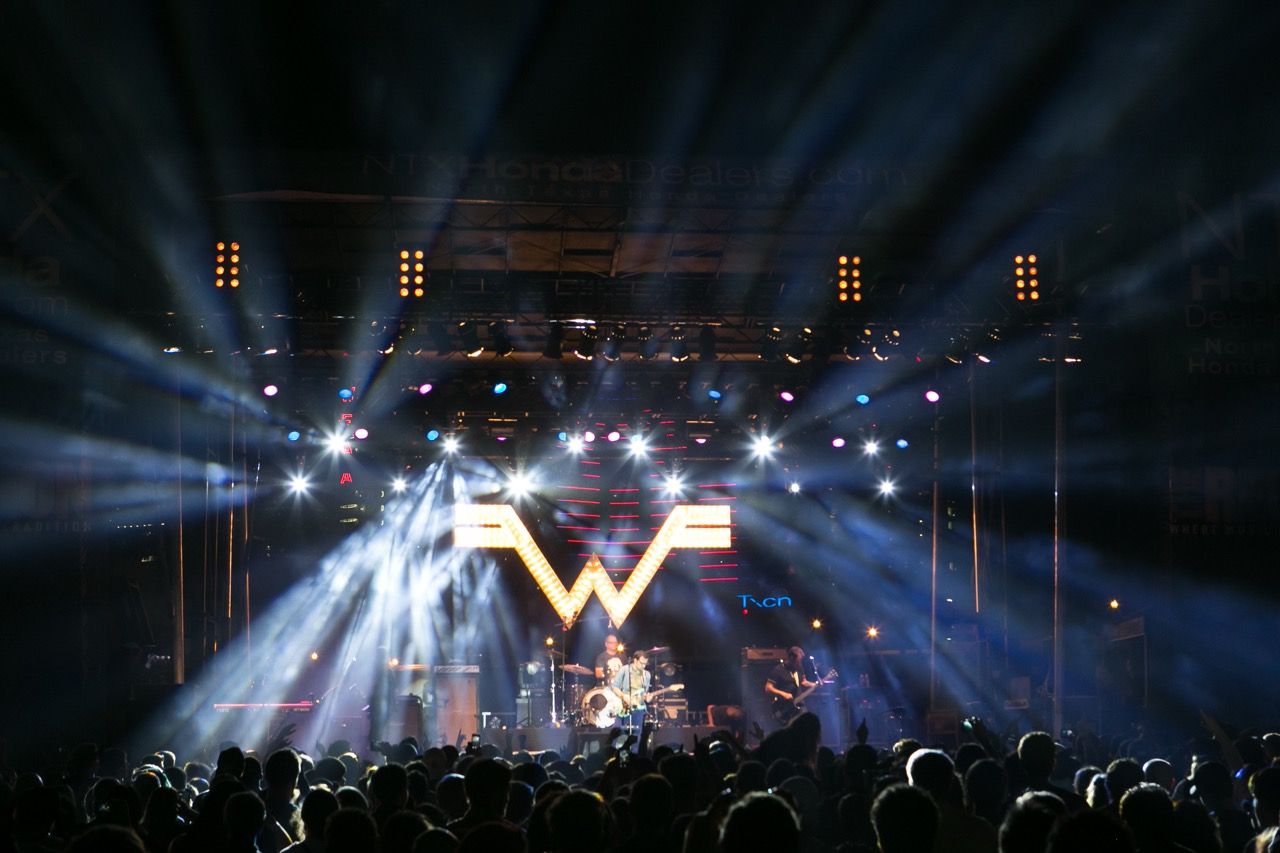 Weezer performing live on stage with dramatic light beams and iconic W logo backdrop before a large crowd.