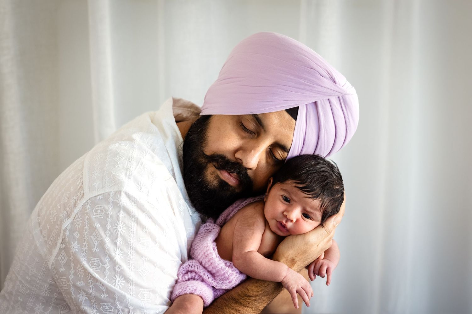 A Sikh father in a pink turban tenderly holds his sleeping newborn baby wrapped in a soft lavender blanket.