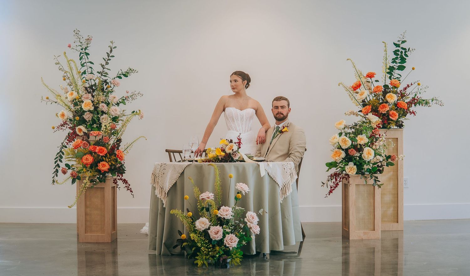 Bride and groom seated at elegant sweetheart table with floral arrangements and warm lighting in modern wedding venue.