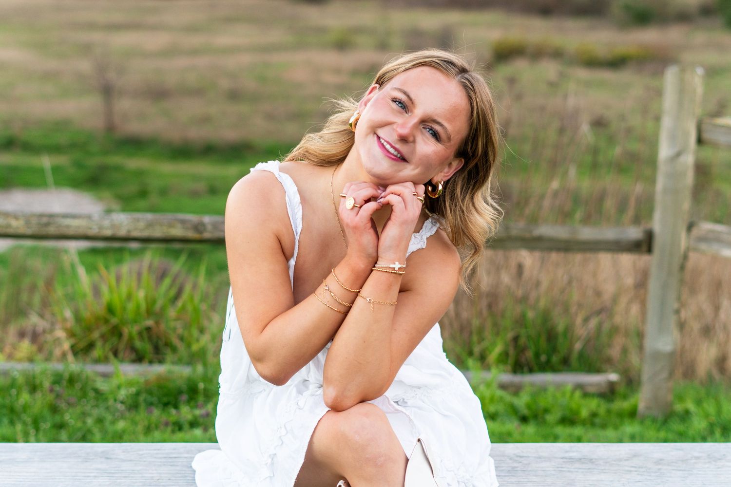 Smiling blonde woman in white dress sitting outdoors near wooden fence with green fields in background.