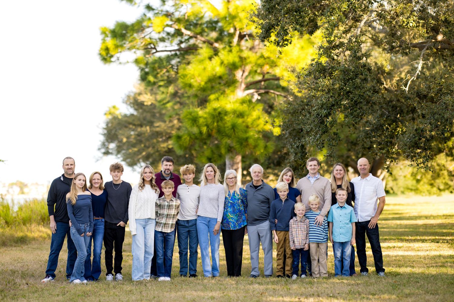Large family group portrait outdoors on green grass with trees in background on a sunny day.