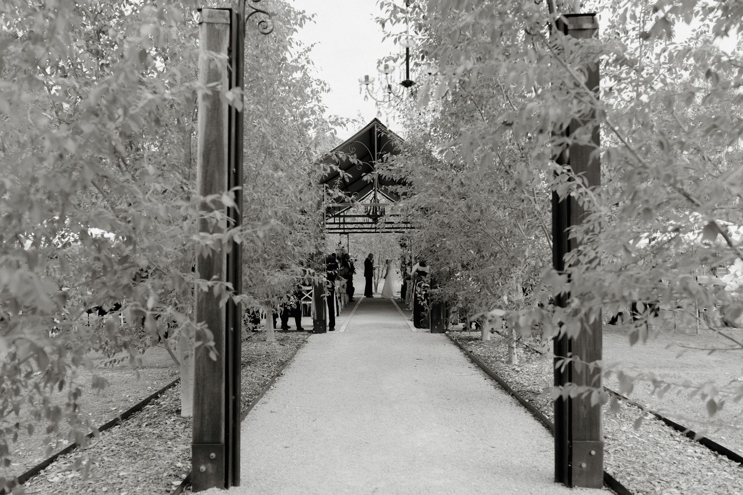 Black and white infrared photo of a tree-lined pathway leading to a chapel with silhouetted figures.