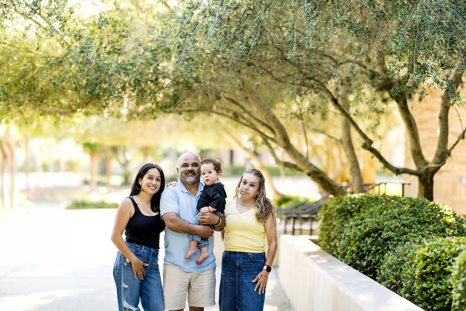 Family of four posing outdoors under olive trees on a sunny day, parents holding a toddler between two women.