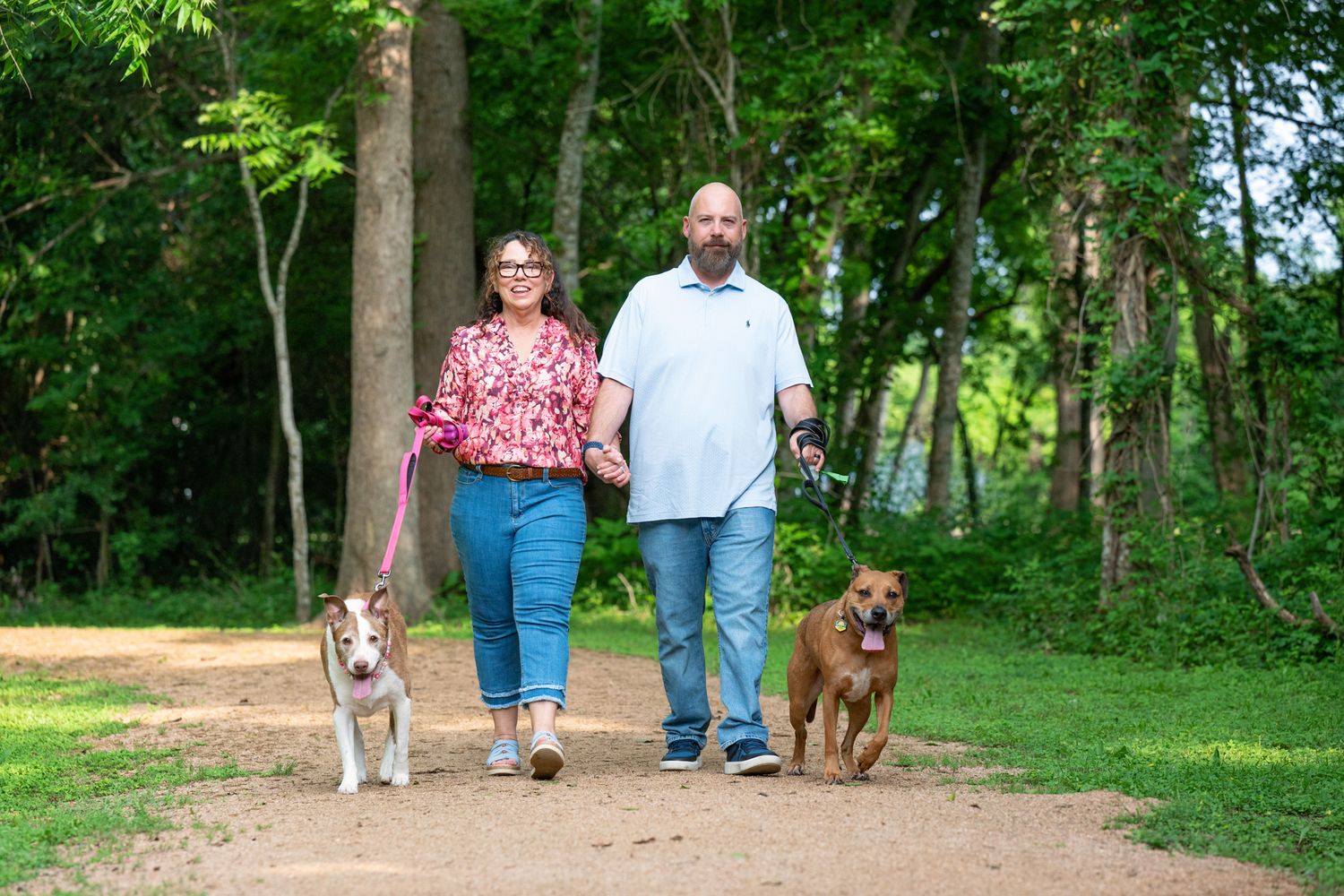 Couple walking two dogs on a tree-lined park path, woman in red floral top and man in white shirt, sunny day.