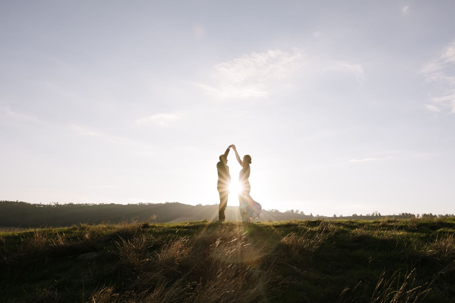 Silhouette of couple dancing in golden sunset light in a scenic field with trees in the background.