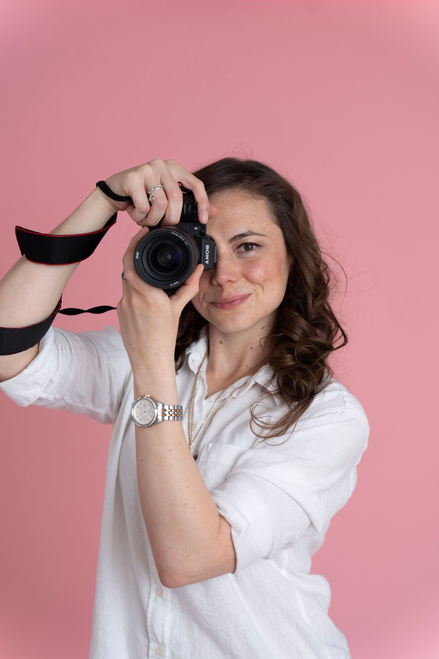Woman in white shirt holding a DSLR camera on her shoulder, posing confidently against a tan background.