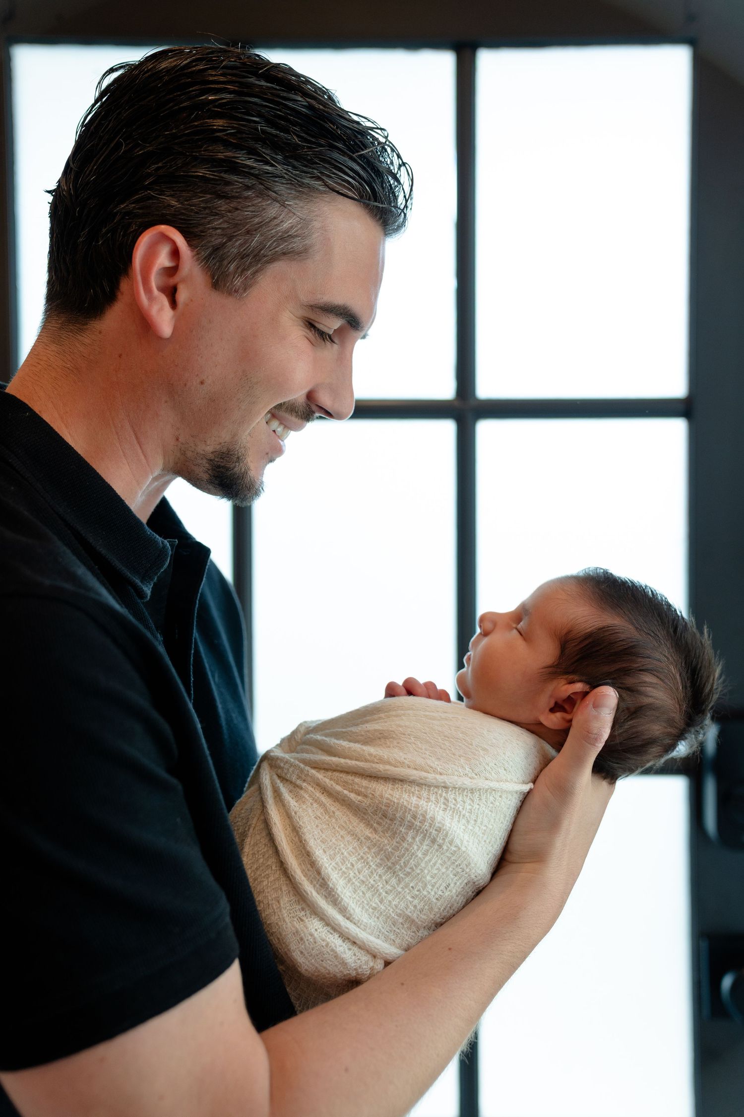 Smiling father in black shirt tenderly holds swaddled newborn baby near bright window.