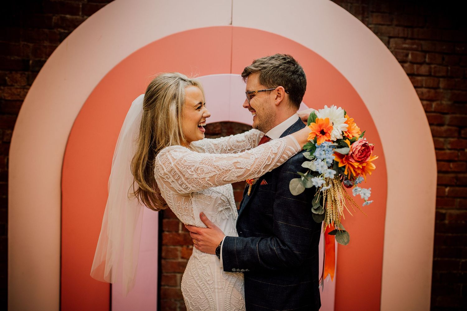 A couple shares intimate moments in front of a coral-colored arched wall during their wedding photoshoot.