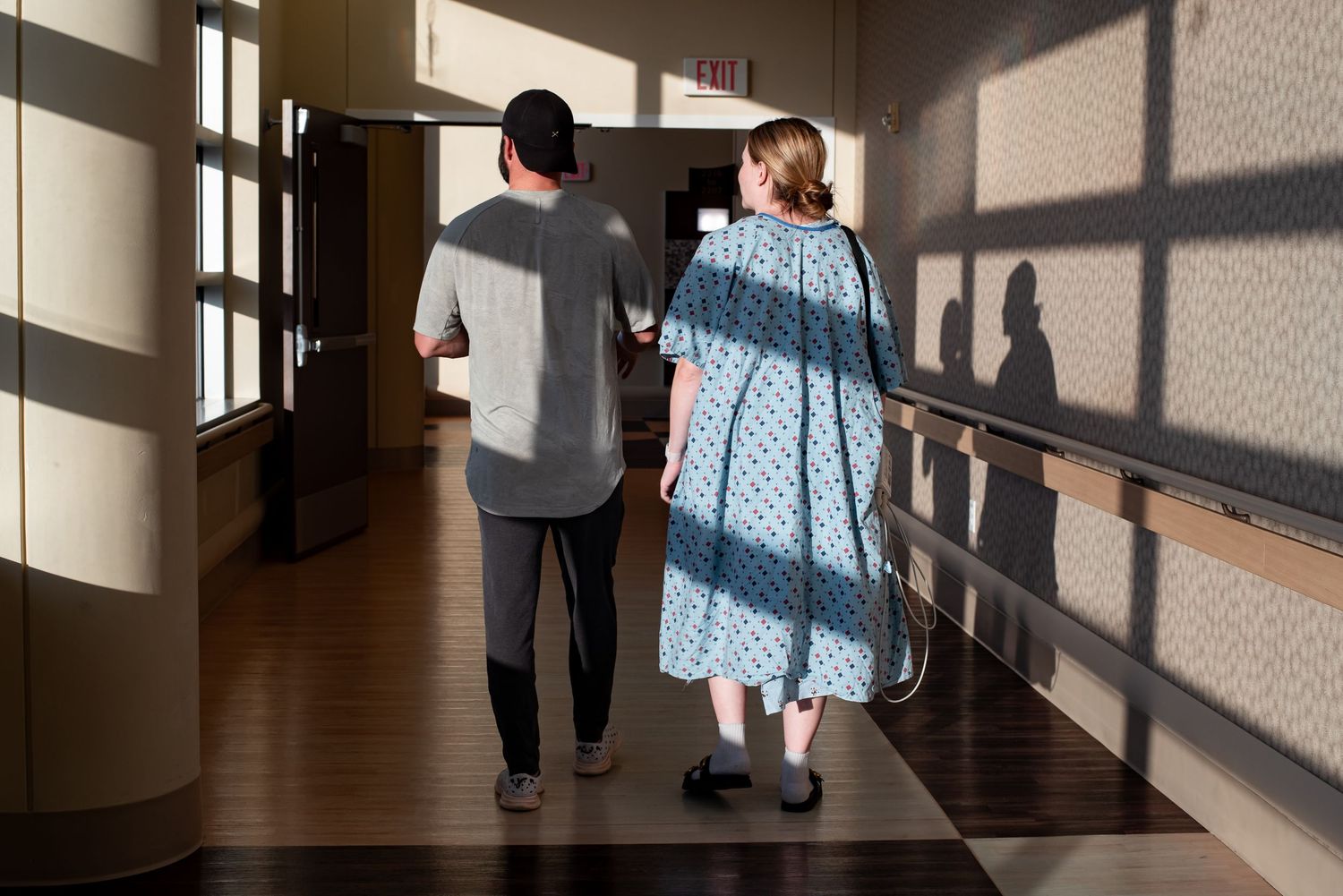 People walking through a sunlit hallway with dramatic shadows cast on the wall from windows.