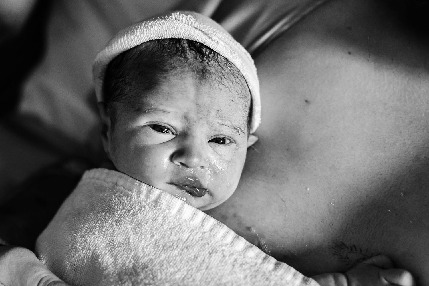 Newborn baby sleeping peacefully while being held skin-to-skin, wearing a white hat, with rosy cheeks and tiny fingers.