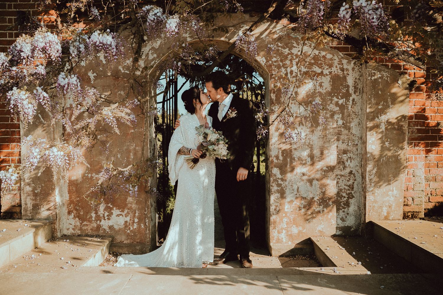 Bride and groom share a romantic moment under a floral arch with wisteria blooms at an elegant wedding venue.