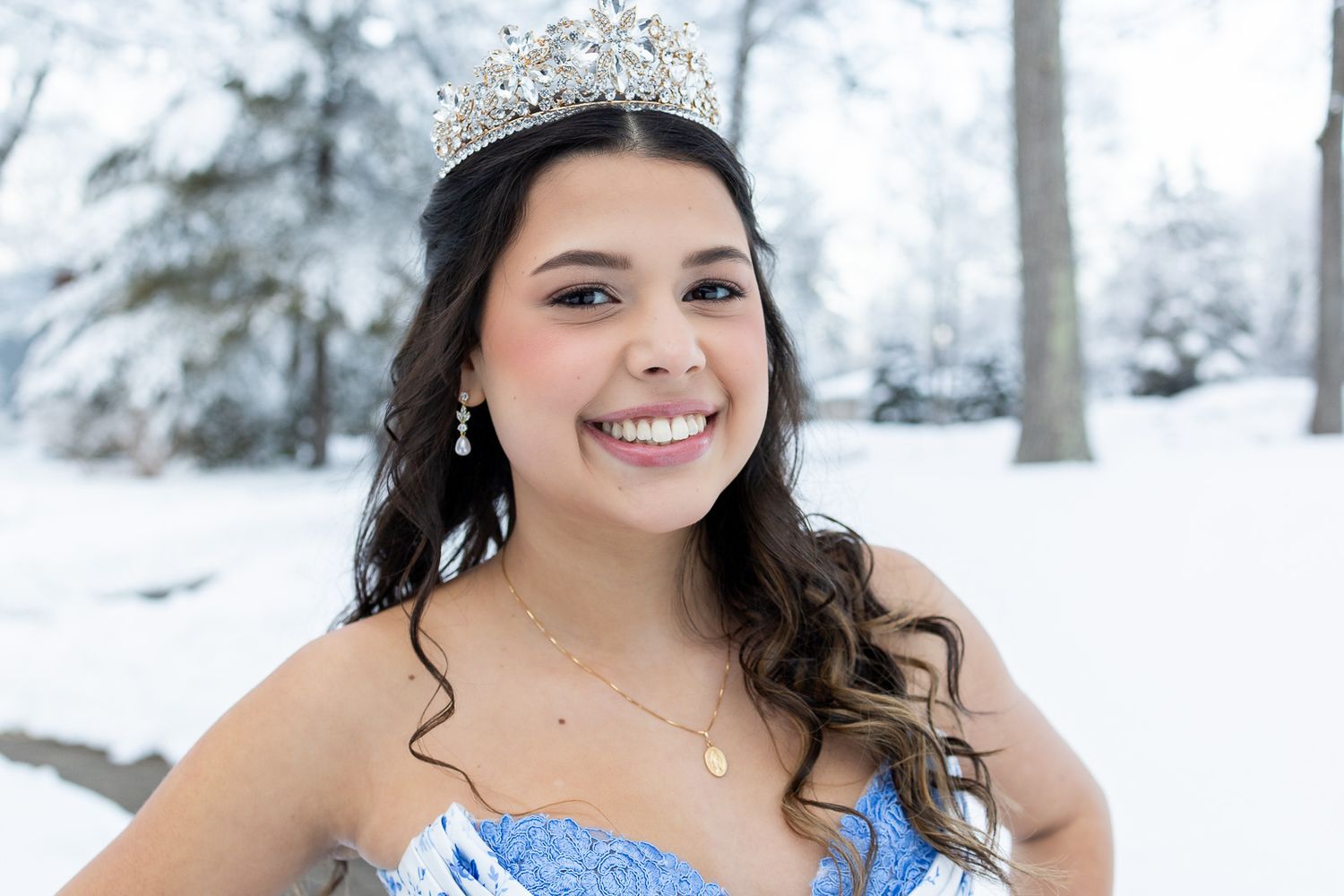 Young woman wearing a sparkling tiara and blue gown smiles outdoors in a snowy winter setting.