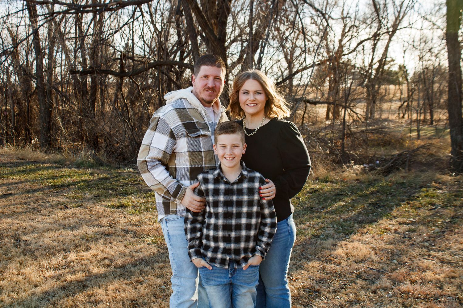 Family of three posing outdoors among bare trees in a golden field during an extended family photo session at The League House in Amarillo