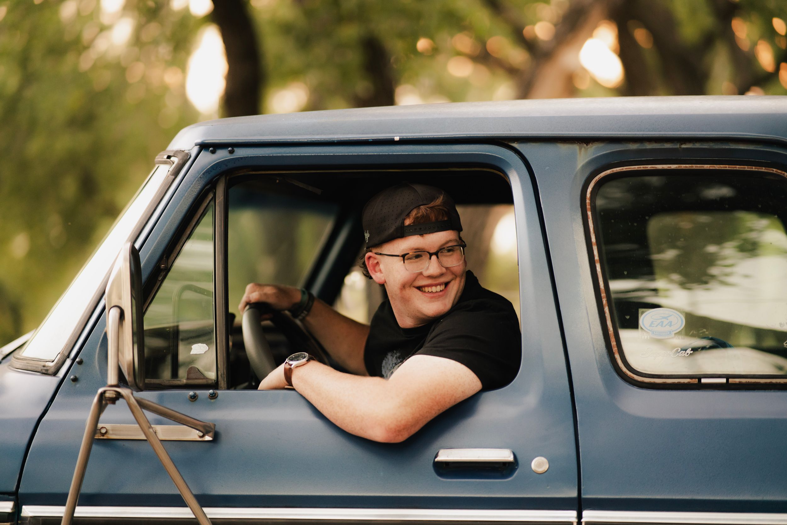 Smiling young man wearing glasses sitting in a vintage blue pickup truck with window down on a sunny day.