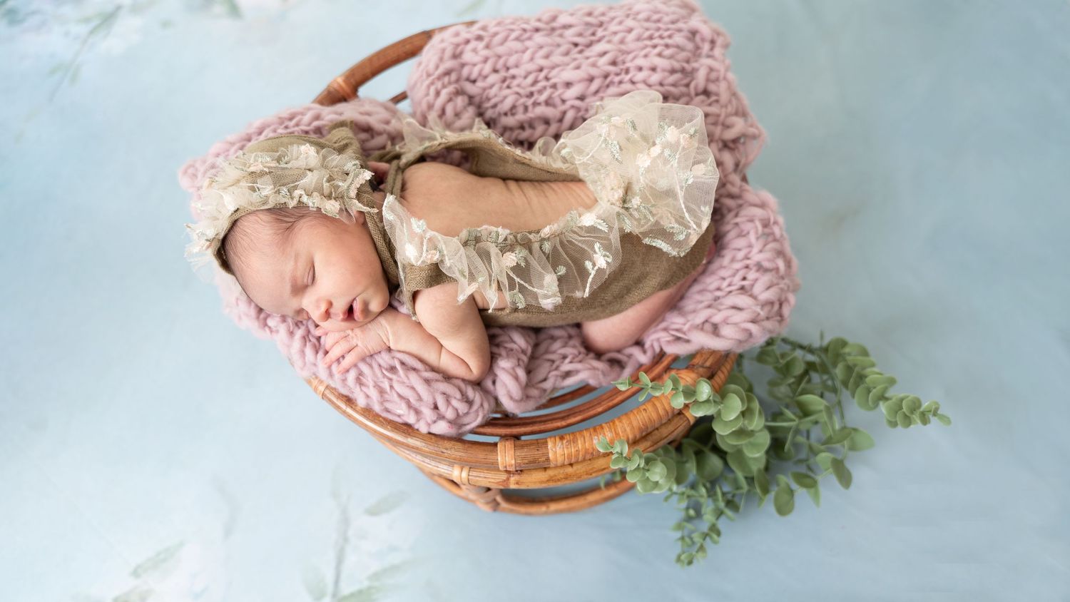 Sleeping baby in pink blanket lying in woven basket with green foliage accent during newborn photo session.