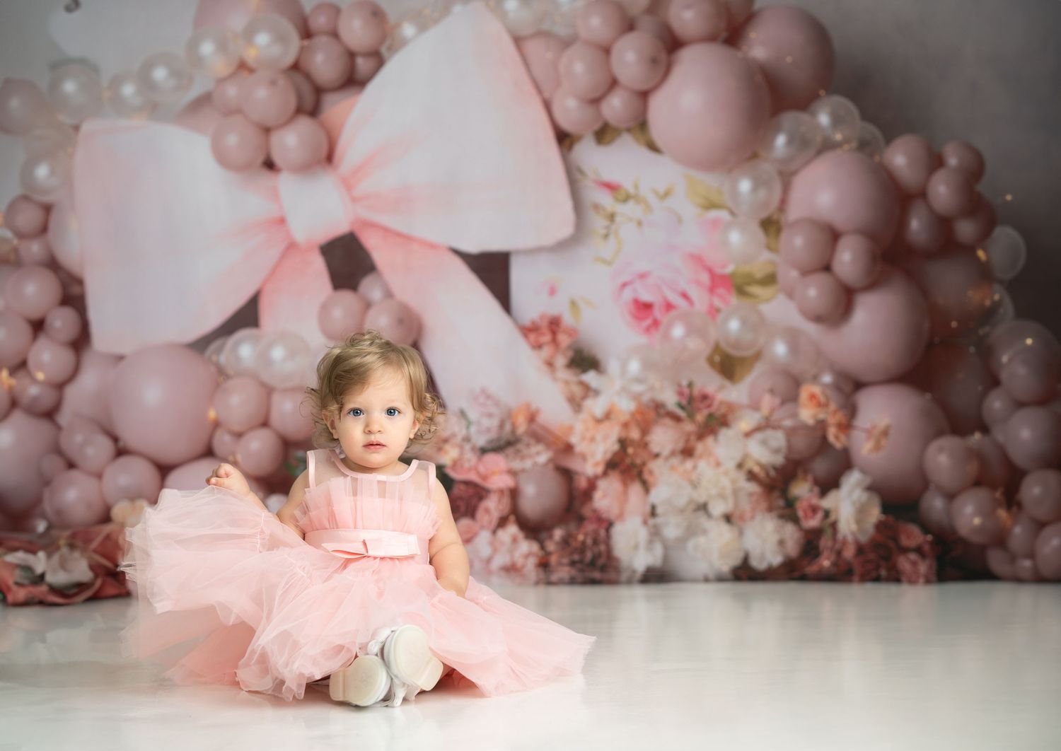A pink balloon arch and floral backdrop creates a dreamy setting with tulle dress and bow decorations.