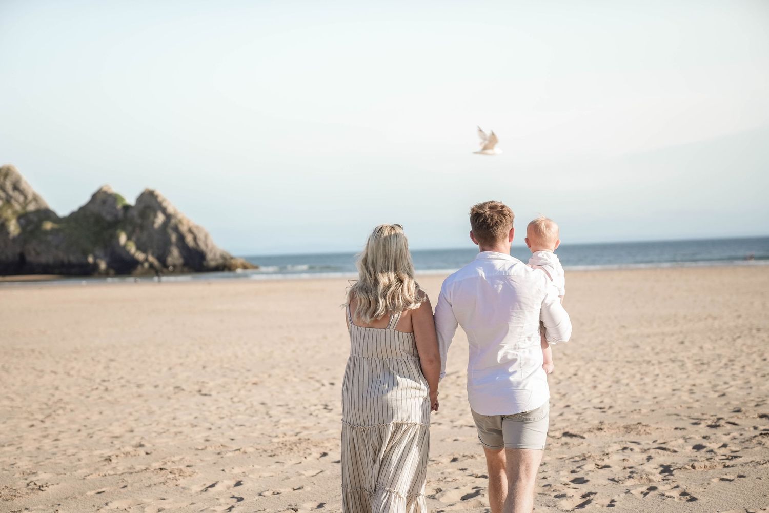 Couple walking along a scenic beach during sunset, holding hands with ocean waves and rocky cliffs in the background.