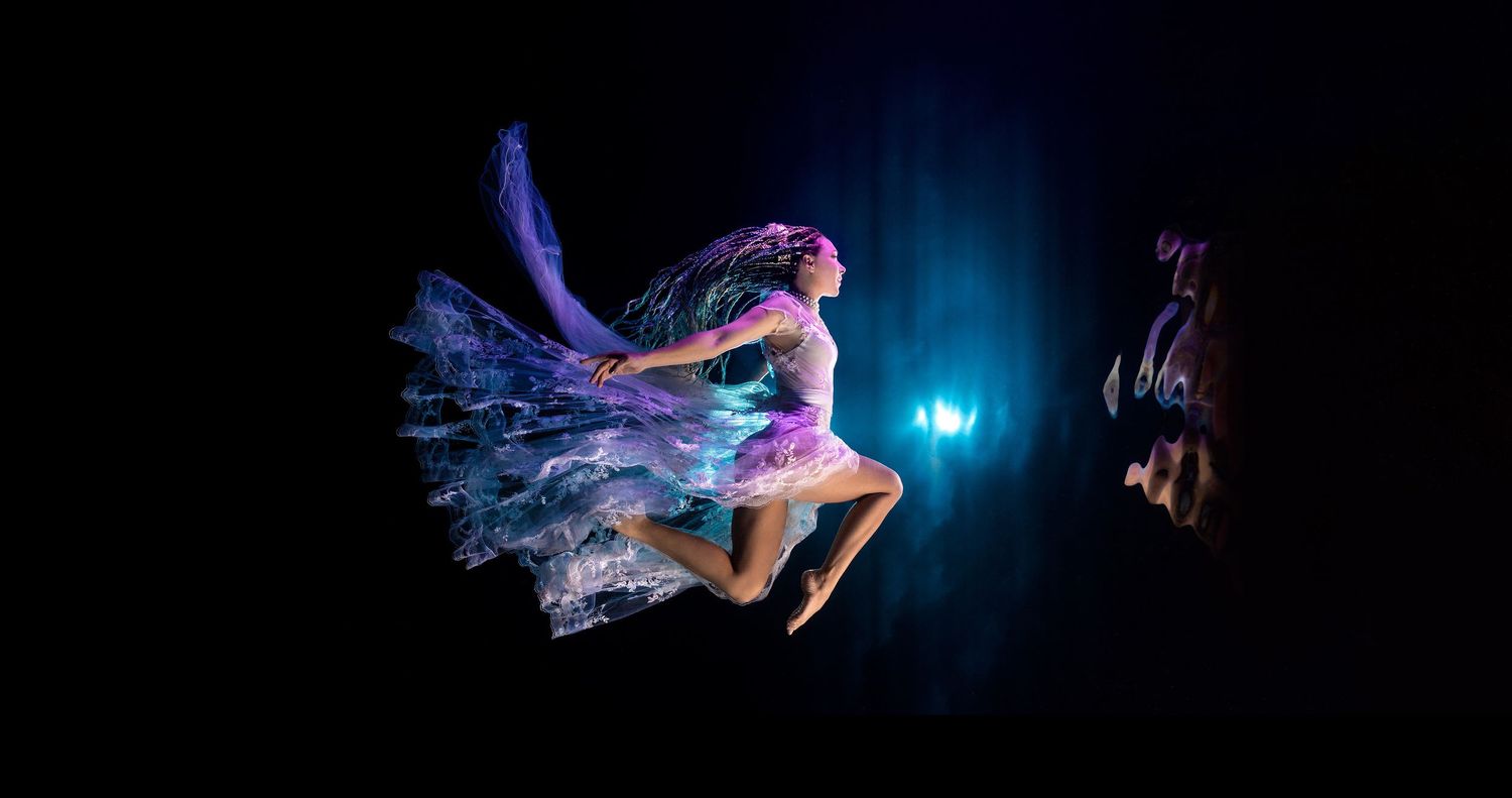 A graceful dancer in flowing fabric performs against a dark background with dramatic blue lighting.