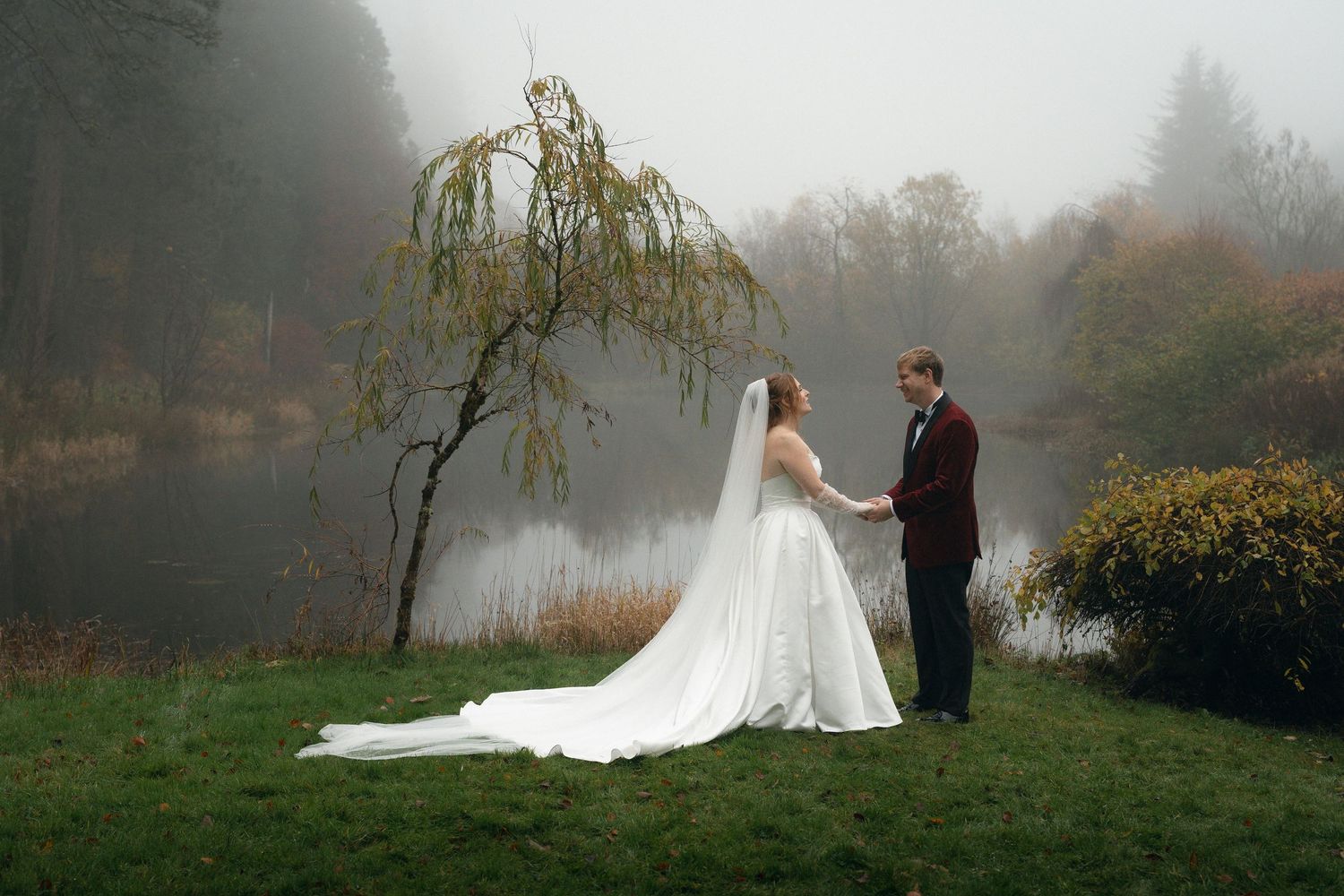 Couple walking through a garden path in wedding attire on a rainy day with umbrellas.