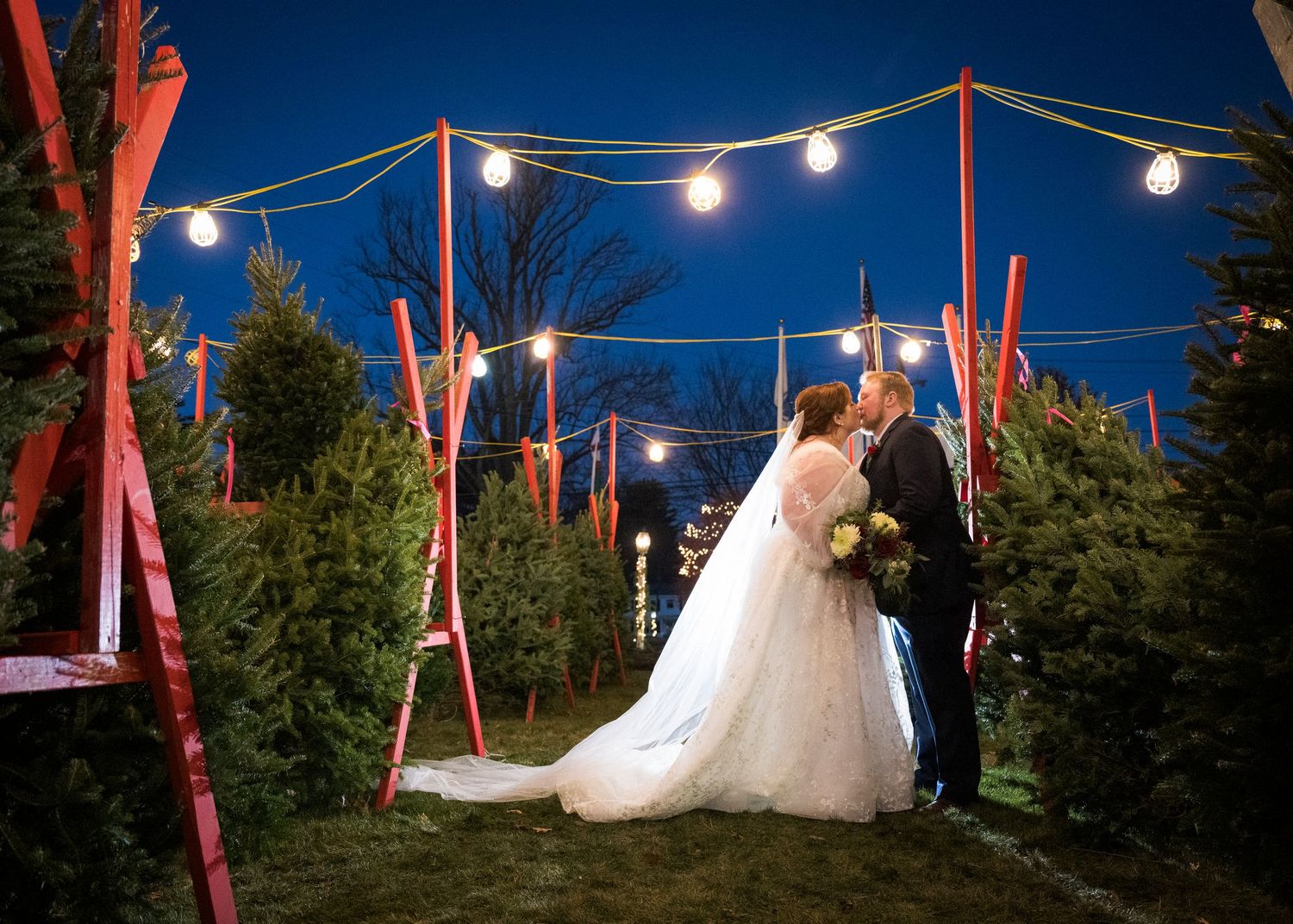 A wedding couple poses together in a field with tall pine trees in the background at sunset.