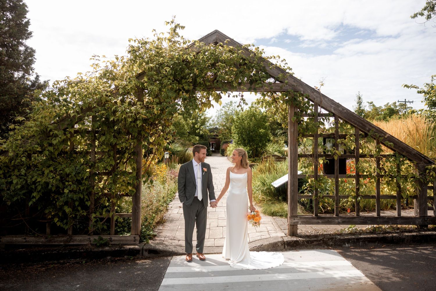 A couple in wedding attire walks together through a rustic outdoor pathway surrounded by lush greenery and natural light.