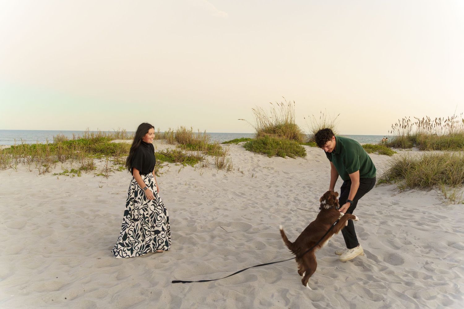 People walking with a dog on a sandy beach path through coastal dunes and vegetation at sunset.