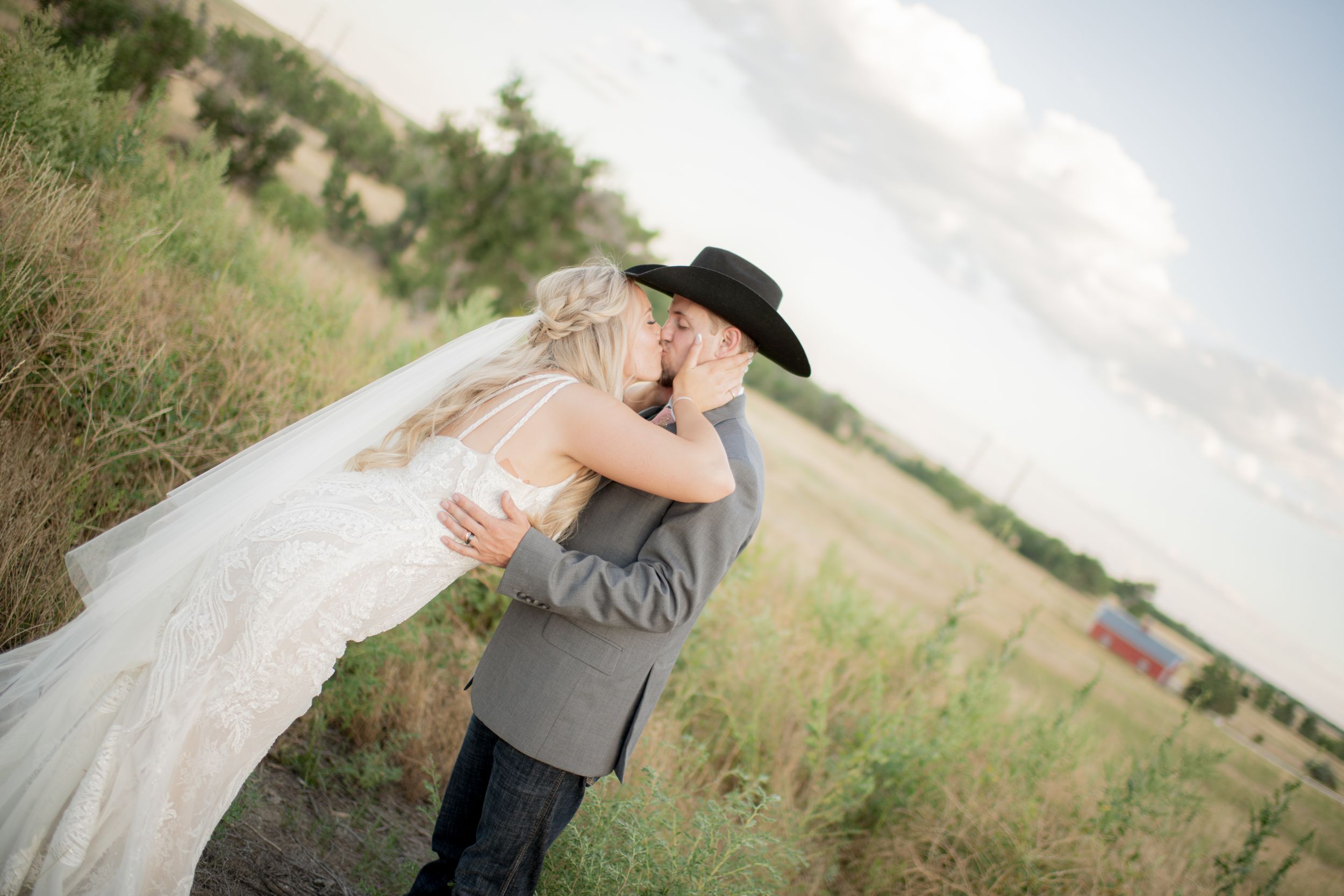 A couple in wedding attire stands together in a sunlit field of tall grass and wildflowers.