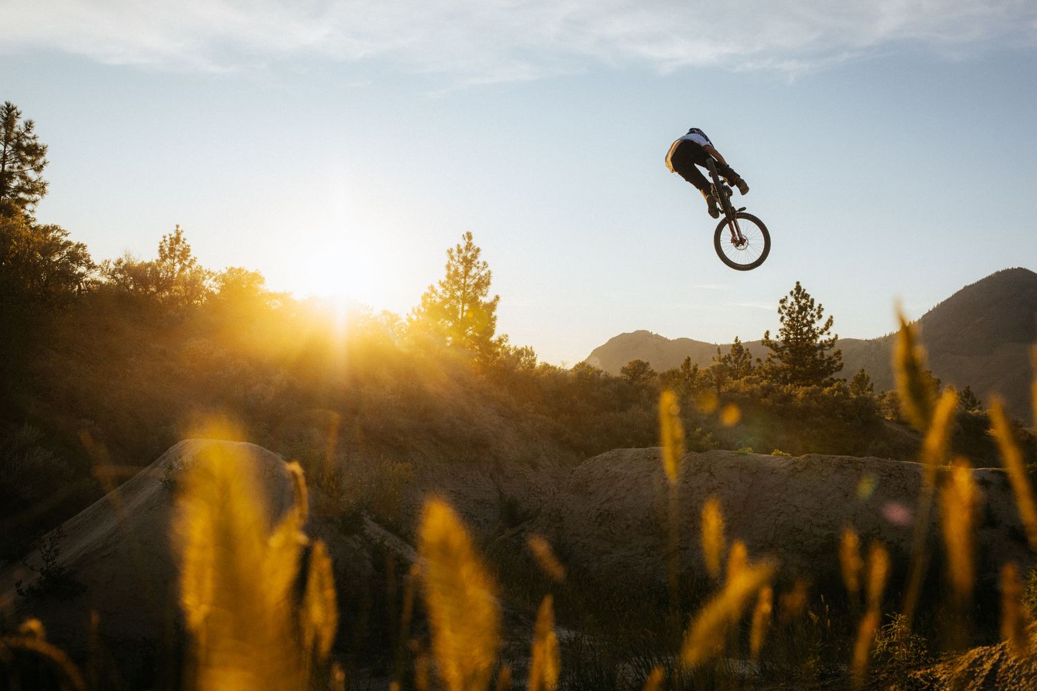 Mountain biker performs aerial trick against golden sunset sky with mountain silhouettes in background.