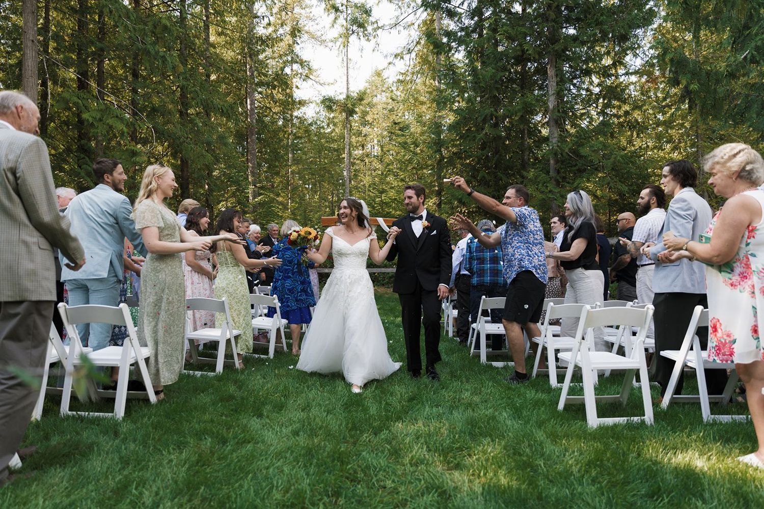Bride and groom walk down outdoor aisle surrounded by cheering guests at a forest wedding ceremony.