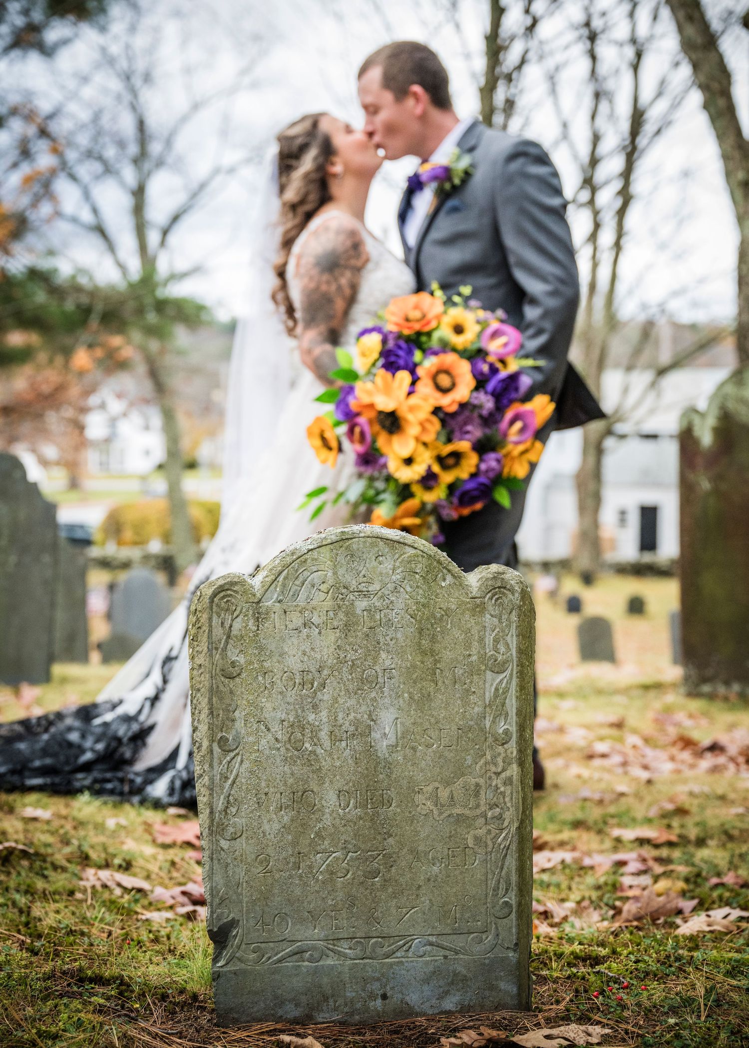 A wedding couple poses together in a field with tall pine trees in the background at sunset.