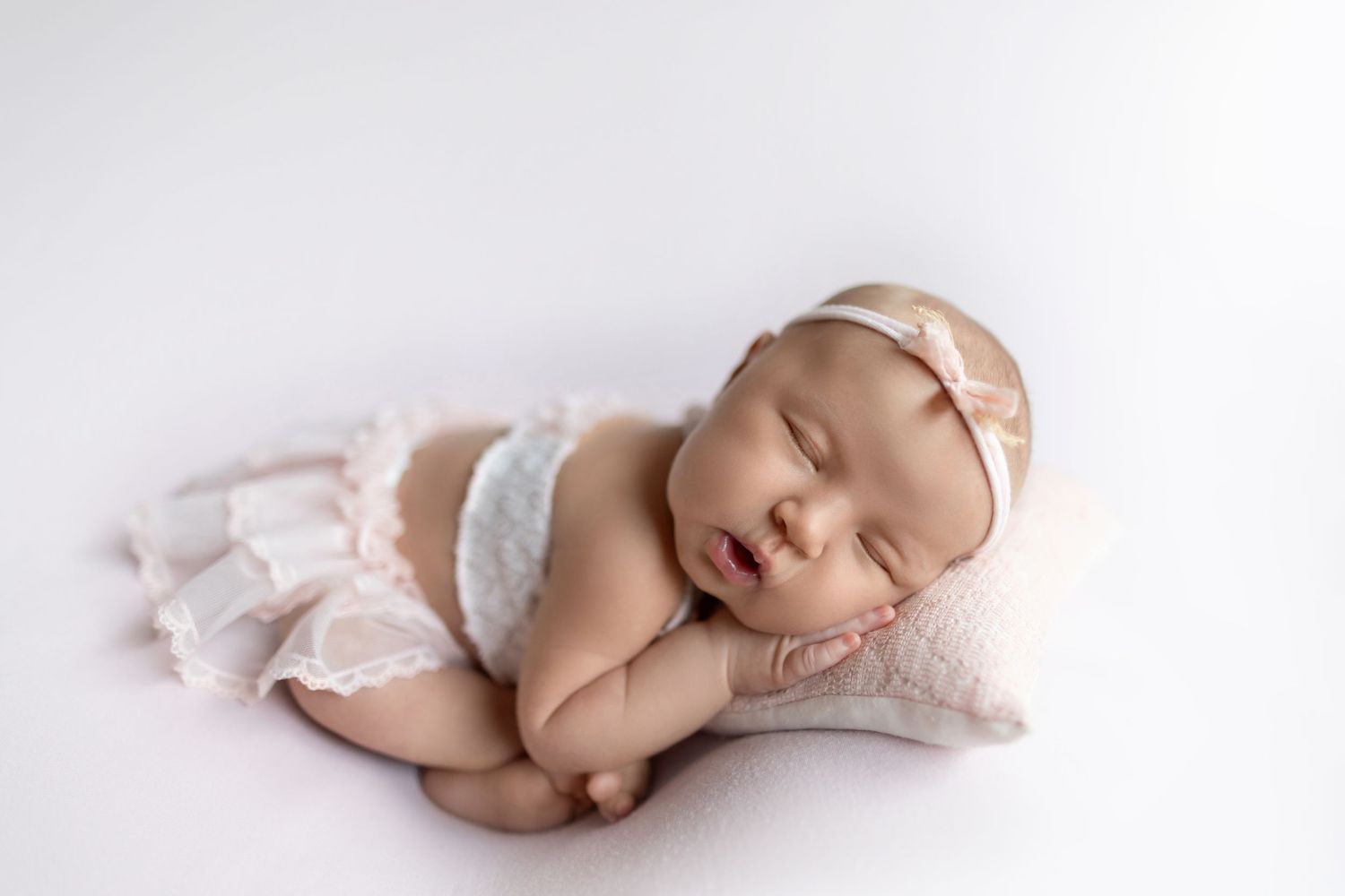 Sleeping newborn baby wearing white lace diaper cover lies peacefully on soft white blanket.
