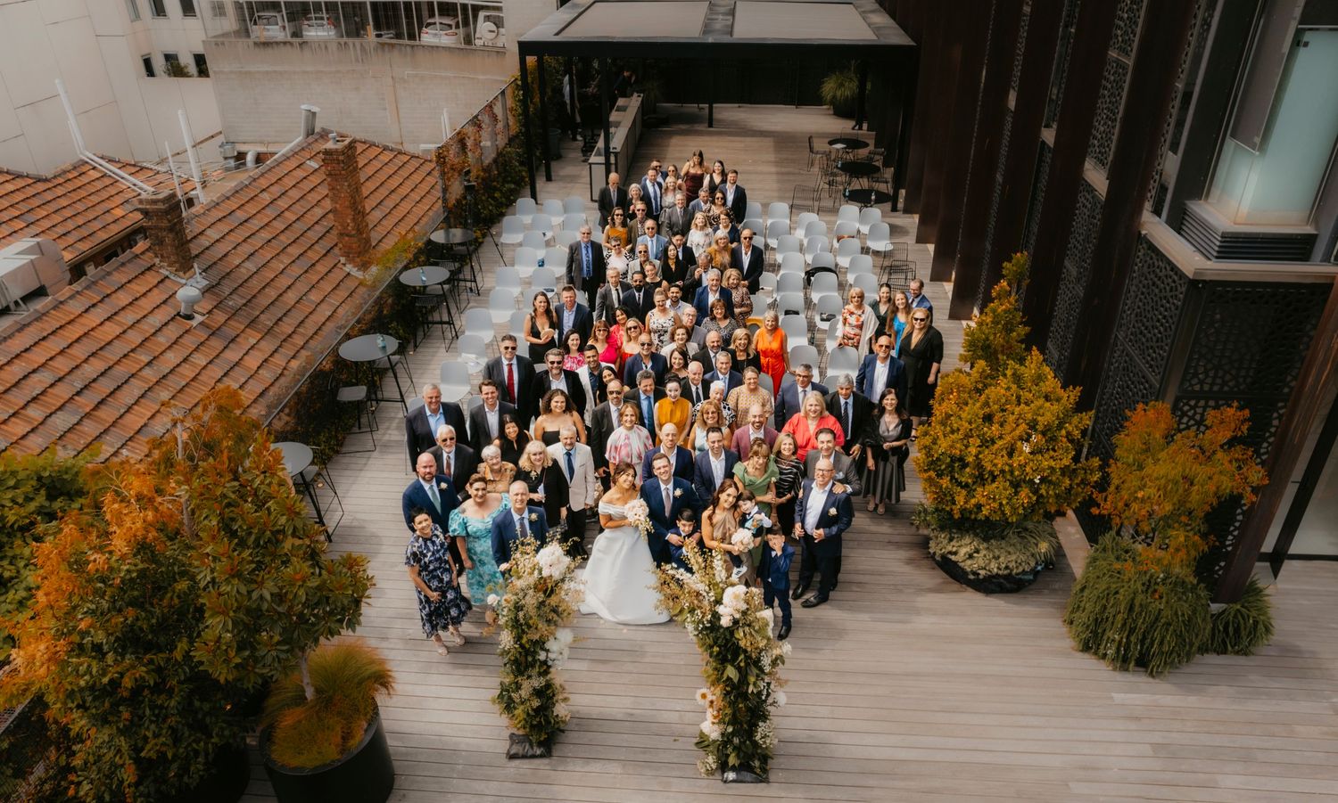 Aerial view of wedding guests gathered on an outdoor rooftop terrace surrounded by autumn foliage and urban buildings.