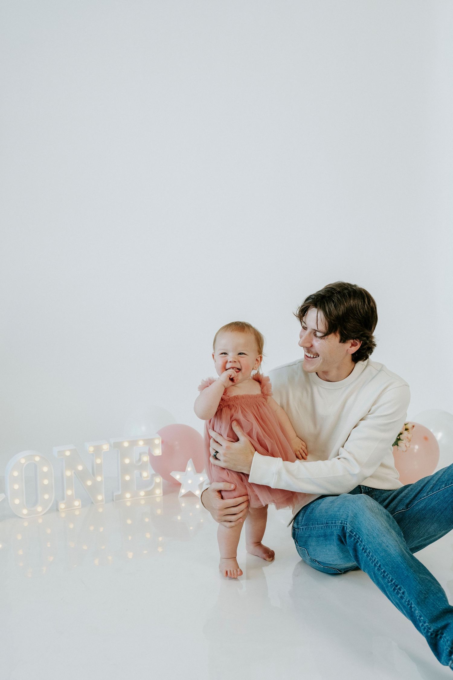 Parent and toddler in pink tulle dress share playful moments on white floor with twinkling lights in background.
